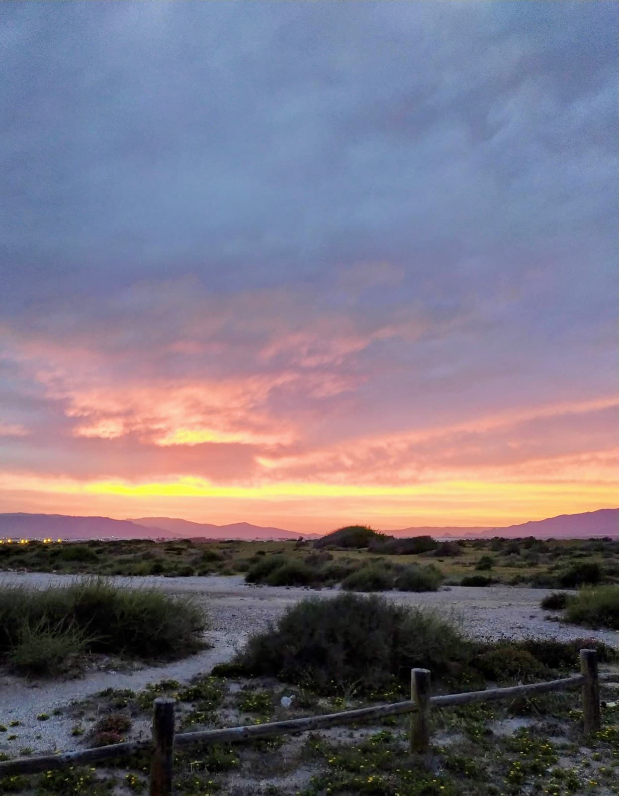 Atardecer sobre una zona de matorral costero, con una valla de madera y montañas en el horizonte